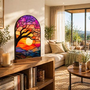 Peut inclure: Lampe en vitrail représentant un coucher de soleil avec une silhouette d'arbre. La lampe, avec un socle en bois, présente un design coloré avec des nuances de violet, d'orange et de jaune. Elle est posée sur une étagère en bois avec des livres et des bougies.