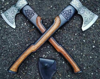 May include: Two crossed axes with silver blades and wooden handles. The axe heads feature intricate designs, including a snowflake and a triangular symbol. The handles are brown with a dotted pattern. A black leather sheath rests below the axes.