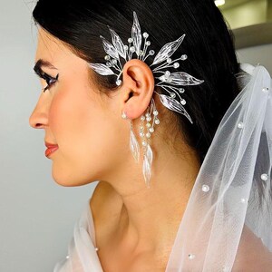 Puede incluir: Un conjunto de pendientes y puño de oreja plateados con un diseño de hojas y cristales. El puño de oreja se envuelve alrededor de la oreja con un diseño floral, y los pendientes cuelgan con colgantes transparentes en forma de hoja y pequeñas perlas. También se ve un velo blanco.