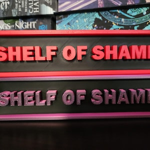 以下が含まれることがあります： 「SHELF OF SHAME」の文字が入った2つの長方形の看板。上の看板は赤いフレームと黒い背景に赤い文字が入っています。下の看板は紫色のフレームと黒い背景に文字が入っています。