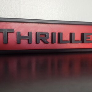 Puede incluir: Un letrero rectangular con la palabra "THRILLER" en letras negras en relieve sobre un fondo rojo. El letrero est&aacute; enmarcado en negro y descansa sobre una superficie oscura.