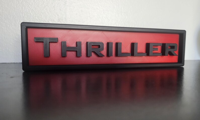 Puede incluir: Un letrero rectangular con la palabra "THRILLER" en letras negras en relieve sobre un fondo rojo. El letrero est&aacute; enmarcado en negro y descansa sobre una superficie oscura.