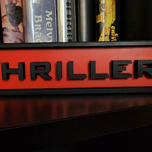 Puede incluir: Un letrero rectangular con la palabra "THRILLER" en letras negras en relieve sobre un fondo rojo. El letrero est&aacute; enmarcado en negro y se encuentra sobre una superficie oscura, con libros visibles en el fondo.