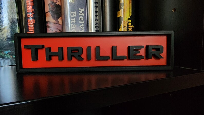 Puede incluir: Un letrero rectangular con la palabra "THRILLER" en letras negras en relieve sobre un fondo rojo. El letrero est&aacute; enmarcado en negro y se encuentra sobre una superficie oscura, con libros visibles en el fondo.