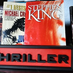 Puede incluir: Una colecci&oacute;n de libros, incluyendo t&iacute;tulos de Stephen King y Michael Crichton, expuestos en una estanter&iacute;a. Un letrero rojo y negro que dice "THRILLER" se encuentra frente a los libros. Los libros son de varios colores, incluyendo rojo, blanco y azul.