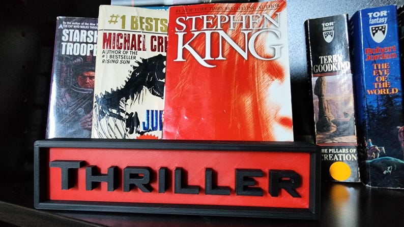 Puede incluir: Una colecci&oacute;n de libros, incluyendo t&iacute;tulos de Stephen King y Michael Crichton, expuestos en una estanter&iacute;a. Un letrero rojo y negro que dice "THRILLER" se encuentra frente a los libros. Los libros son de varios colores, incluyendo rojo, blanco y azul.