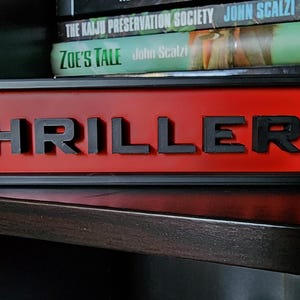 Puede incluir: Un letrero rectangular con la palabra "THRILLER" en letras negras en relieve sobre un fondo rojo. El letrero est&aacute; enmarcado en negro y se encuentra en una estanter&iacute;a oscura, con libros en el fondo.