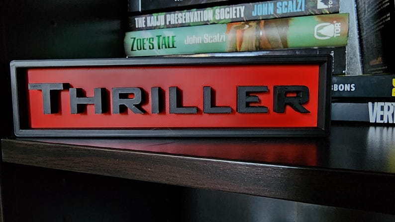 Puede incluir: Un letrero rectangular con la palabra "THRILLER" en letras negras en relieve sobre un fondo rojo. El letrero est&aacute; enmarcado en negro y se encuentra en una estanter&iacute;a oscura, con libros en el fondo.