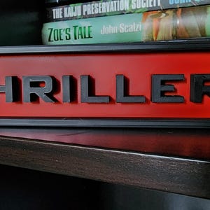 Puede incluir: Un letrero rectangular con la palabra "THRILLER" en letras negras en relieve sobre un fondo rojo. El letrero est&aacute; enmarcado en negro y descansa sobre una superficie oscura, con libros visibles en el fondo.