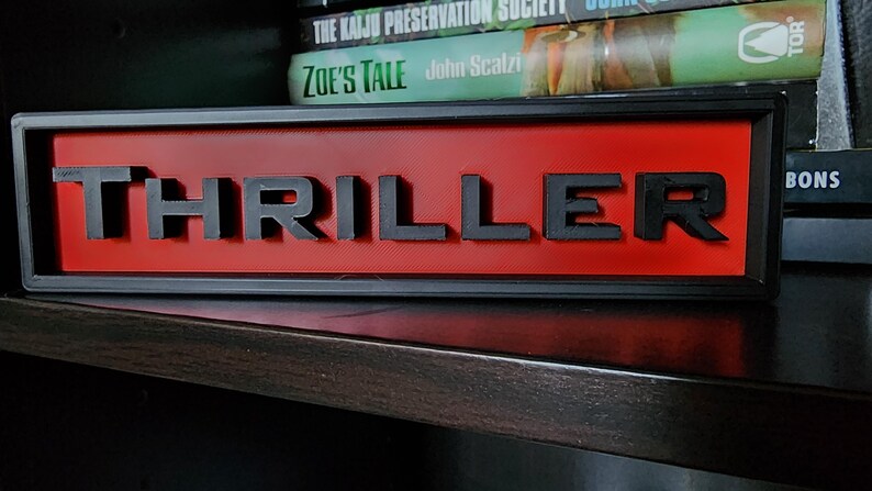 Puede incluir: Un letrero rectangular con la palabra "THRILLER" en letras negras en relieve sobre un fondo rojo. El letrero est&aacute; enmarcado en negro y descansa sobre una superficie oscura, con libros visibles en el fondo.