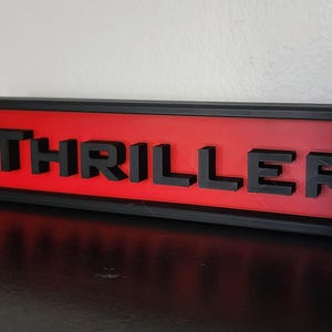Puede incluir: Un letrero rectangular con la palabra "THRILLER" en letras negras en relieve. Las letras est&aacute;n sobre un fondo rojo, enmarcadas por un borde negro. El letrero est&aacute; sobre una superficie oscura.
