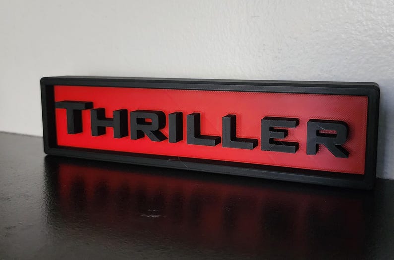 Puede incluir: Un letrero rectangular con la palabra "THRILLER" en letras negras en relieve. Las letras est&aacute;n sobre un fondo rojo, enmarcadas por un borde negro. El letrero est&aacute; sobre una superficie oscura.