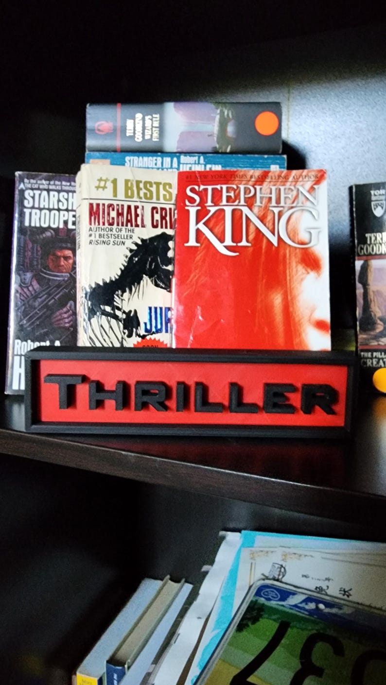 Puede incluir: Una estanter&iacute;a muestra varios libros, incluyendo t&iacute;tulos de Stephen King y Michael Crichton. Un letrero rojo y negro que dice "THRILLER" se encuentra frente a los libros, indicando el g&eacute;nero. Los libros est&aacute;n apilados en una estanter&iacute;a de madera oscura.