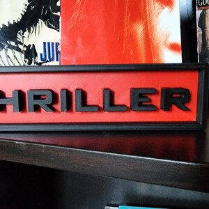 Puede incluir: Un letrero rectangular con la palabra "THRILLER" en letras negras en relieve. El fondo es rojo y el letrero est&aacute; enmarcado en negro. El letrero se muestra en una estanter&iacute;a oscura, con libros al fondo.