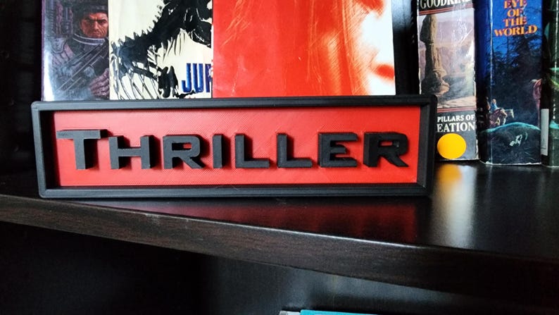 Puede incluir: Un letrero rectangular con la palabra "THRILLER" en letras negras en relieve. El fondo es rojo y el letrero est&aacute; enmarcado en negro. El letrero se muestra en una estanter&iacute;a oscura, con libros al fondo.