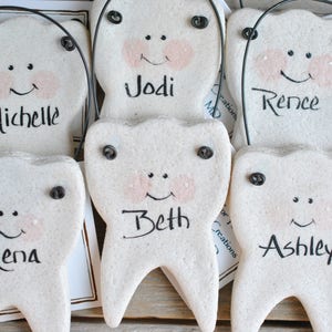 May include: Five white ceramic tooth ornaments with black eyes and mouths, each with a different name written on them: Jodi, Michelle, Rence, Beth, and Ashley.
