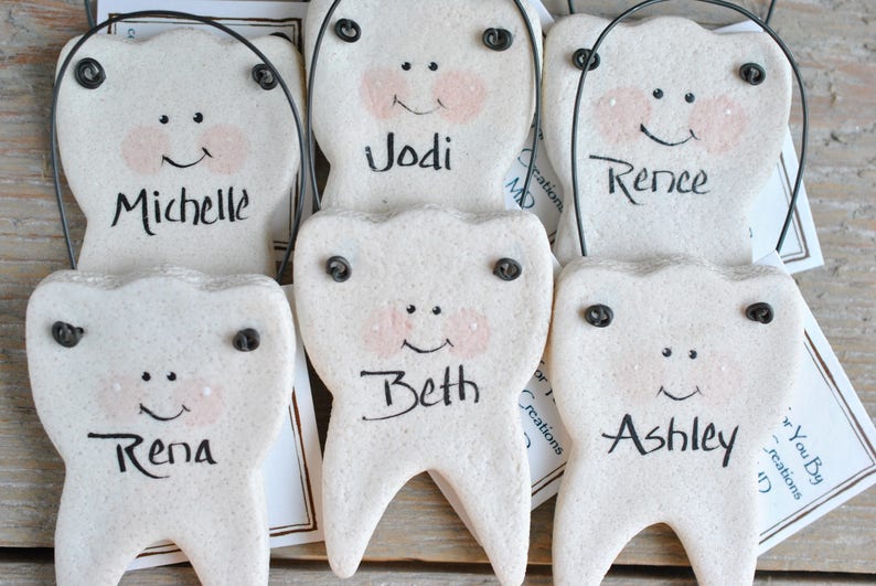May include: Five white ceramic tooth ornaments with black eyes and mouths, each with a different name written on them: Jodi, Michelle, Rence, Beth, and Ashley.