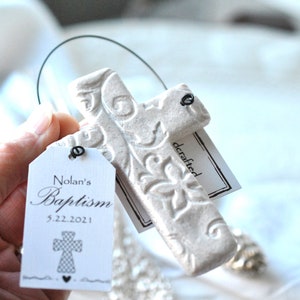 Puede incluir: Cruz de cer&aacute;mica blanca con un dise&ntilde;o floral en relieve, colgada de un lazo de alambre. Una etiqueta blanca con texto negro dice "Nolan's Baptism 5.22.2021" y un peque&ntilde;o gr&aacute;fico de cruz. Una segunda etiqueta dice "crafted".