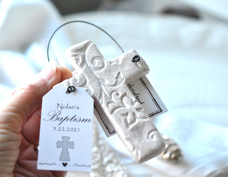 Puede incluir: Cruz de cer&aacute;mica blanca con un dise&ntilde;o floral en relieve, colgada de un lazo de alambre. Una etiqueta blanca con texto negro dice "Nolan's Baptism 5.22.2021" y un peque&ntilde;o gr&aacute;fico de cruz. Una segunda etiqueta dice "crafted".