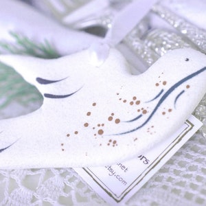 May include: White ceramic dove ornament with brown and blue painted details. The dove has a stylized wing design and brown dots scattered across its body. The ornament is sitting on a white crocheted surface with a small white tag that reads "etsy.com - .net ARTS".