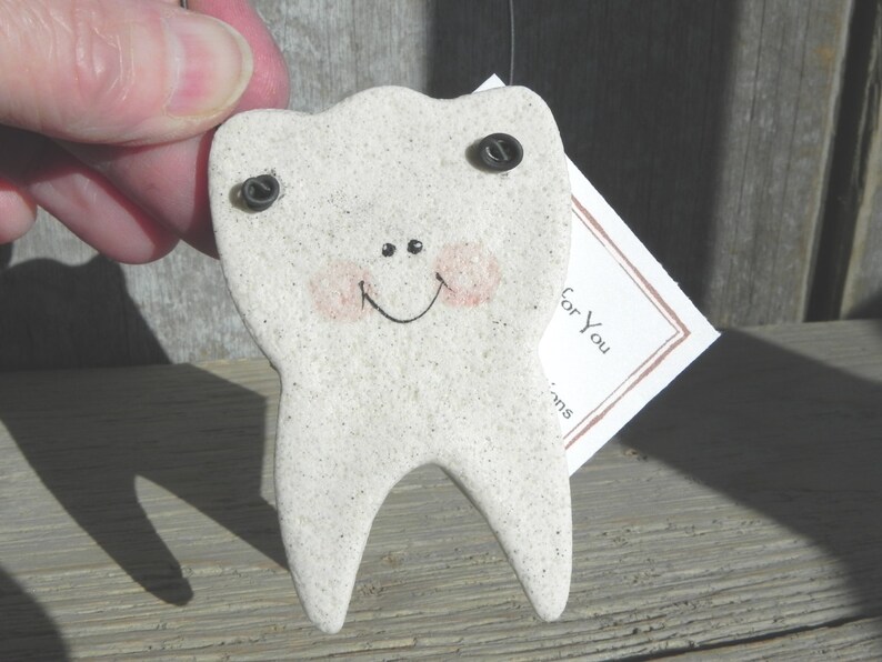 May include: A white ceramic tooth with a smiling face, painted with pink cheeks and black eyes. The tooth has two black screws for eyes and is attached to a small white tag that says "For You".