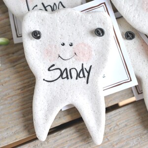 May include: A white ceramic tooth with a smiling face and the name "Sandy" written on it. The tooth has two black buttons for eyes.
