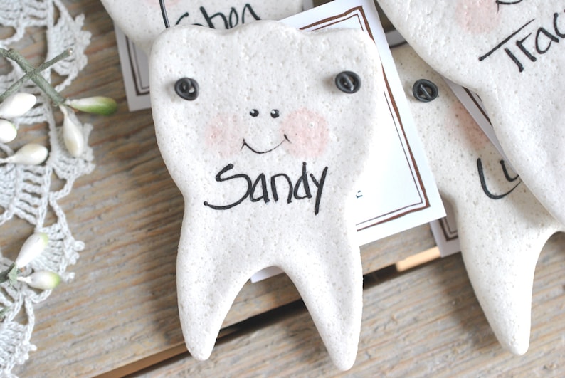 May include: A white ceramic tooth with a smiling face and the name "Sandy" written on it. The tooth has two black buttons for eyes.