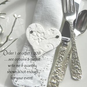 May include: White ceramic heart-shaped napkin ring with a delicate floral design. The napkin ring is on a white linen napkin with silver cutlery. The text on the image reads: "Order 1 or order 1,000! ...see options & please write me if quantity shown is not enough for your event!"