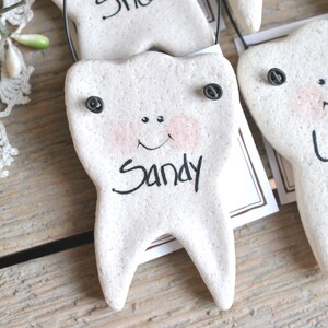 May include: Three white ceramic tooth ornaments with black eyes and painted smiles. Each tooth has a black string for hanging and a name written on it: Sandy, Lucy, and another name that is unclear.