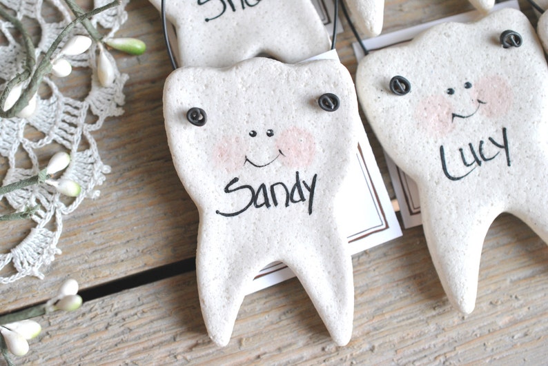 May include: Three white ceramic tooth ornaments with black eyes and painted smiles. Each tooth has a black string for hanging and a name written on it: Sandy, Lucy, and another name that is unclear.