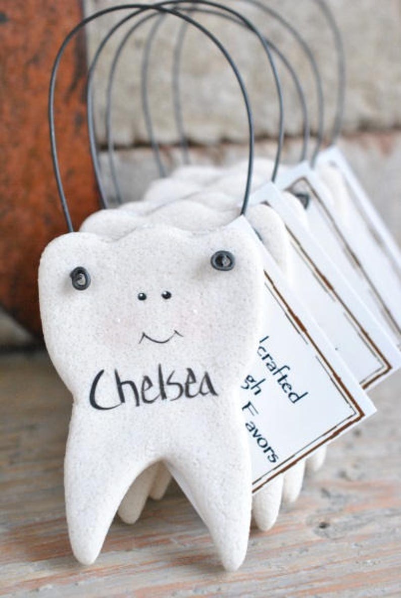 May include: White ceramic tooth ornaments with black eyes and a smile, each with a name written on it. The ornaments are strung on black wire and have a tag that says "I crafted these favors".