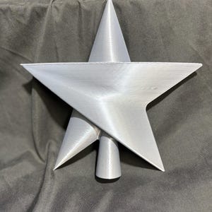 May include: A silver-colored star-shaped Christmas tree topper. The topper has a five-pointed star design with a conical base. The star is made of a metallic material and has a textured surface. The overall design is modern and minimalist.