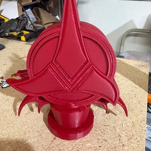 Puede incluir: Una escultura roja vibrante, impresa en 3D, que presenta un emblema estilizado y puntiagudo sobre una base circular. El diseño incluye ángulos agudos y elementos curvos, lo que sugiere un tema futurista o de fantasía. La escultura se exhibe sobre una superficie marrón claro.