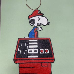 May include: A red, white, and blue Snoopy ornament dressed as Mario, standing on a red doghouse-shaped controller. The controller has a black top with white buttons and a red base. A red string is attached for hanging.