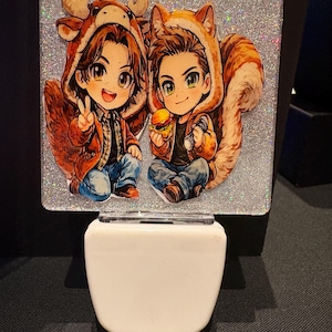 Puede incluir: Luz de noche decorativa con un fondo brillante y una ilustración de dibujos animados de dos figuras con disfraces de animales. Una figura lleva un disfraz de alce, la otra un disfraz de ardilla. La luz de noche tiene una base blanca.