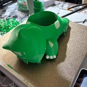Puede incluir: Una maceta verde impresa en 3D con forma de Bulbasaur. La maceta tiene detalles blancos y una gran abertura para plantas. El Bulbasaur está tumbado sobre una superficie dorada.