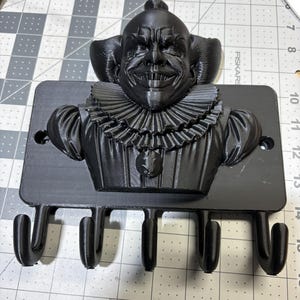 Könnte beinhalten: Ein schwarzer, 3D-gedruckter Wand-Schlüsselhalter mit einem bedrohlichen Clowngesicht mit Rüschenkragen und vier Haken darunter. Der Clown hat ein finsteres Grinsen und einen kleinen Anhänger. Der Schlüsselhalter ist auf einer rechteckigen Basis montiert.