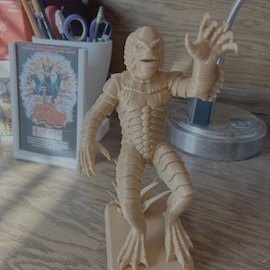 May include: A tan-colored figurine of the Creature from the Black Lagoon, posed with one arm raised. The figure has detailed scales and webbed feet, standing on a rectangular base. The figurine is a collectible item.