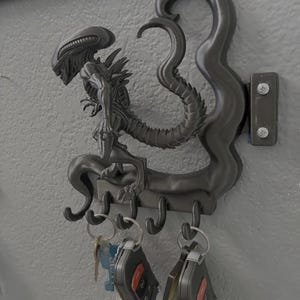 Könnte beinhalten: Ein dunkelgrauer Schlüsselhalter im Alien-Design, der an einer Wand befestigt ist. Der Schlüsselhalter zeigt eine detaillierte Skulptur einer Alien-Kreatur mit vier Haken darunter. Mehrere Schlüsselbunde hängen an den Haken.