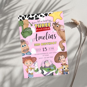 Puede incluir: Una tarjeta de invitaci&oacute;n de cumplea&ntilde;os rosa con personajes de dibujos animados que incluyen un dinosaurio verde, un caballo marr&oacute;n, una figura de patata, una vaquera, un guardi&aacute;n espacial y un vaquero. El texto dice You've Got a Friend Who's THREE, Amelia's 3RD BIRTHDAY, July 15 6 PM.