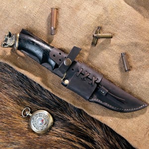 Può includere: Un coltello da caccia con manico nero e fodero in pelle. Il manico presenta un disegno a testa di lupo. Il fodero è fissato con un cinturino e lacci. Sono presenti anche un orologio da tasca e bossoli.