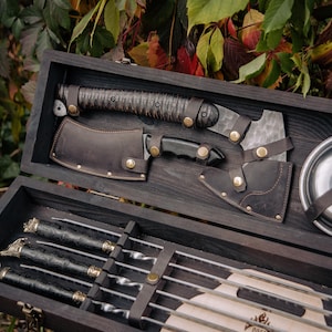 May include: A dark wooden box set containing a hatchet, knife, skewers, and a metal plate with leather straps. The hatchet and knife have leather sheaths. The skewers have decorative handles.