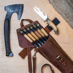May include: Axe with a textured black handle and a dark metal head. A leather quiver holds arrows with brass tips and black and brown shafts. A knife with a decorative handle and a bottle opener is also present.
