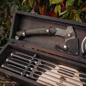 Puede incluir: Un juego de caja de madera oscura que contiene un hacha pequeña con una funda de cuero, brochetas y un plato metálico redondo. El hacha tiene un mango texturizado y una hoja de metal. Las brochetas tienen mangos oscuros. El juego se exhibe al aire libre.