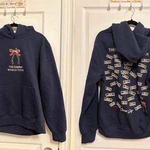 May include: Navy blue hoodie with a front pocket and hood. The front features a red bow and pliers graphic with the text "THE HUSTLER WORLD TOUR". The back has a cassette tape design and text.