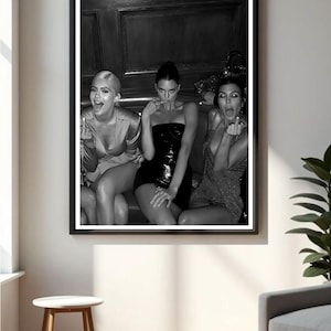 Peut inclure: Impression en noir et blanc encadrée représentant trois femmes. L'une porte une robe bustier noire, les autres des hauts scintillants. Les femmes font des gestes espiègles. L'impression est affichée sur un mur blanc.