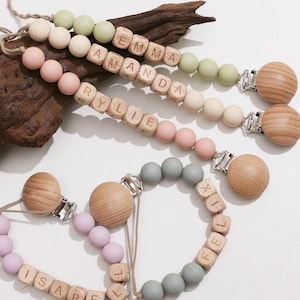 Puede incluir: Pinzas para chupete con cuentas de madera y bloques de nombres personalizados. Las pinzas presentan cuentas de silicona de color pastel en rosa, verde y gris. Los nombres "EMMA", "AMANDA", "RYLIE", "ISABEL" y "FELIX" están escritos en los bloques de madera.