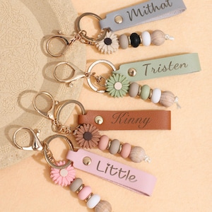 Op de afbeelding: Een verzameling gepersonaliseerde sleutelhangers met namen als "Mithat", "Tristen", "Kinny" en "Little" op leren bandjes. Elke sleutelhanger heeft een bloemvormige bedel en een kralenaccent in verschillende kleuren, bevestigd aan een goudkleurige sluiting.