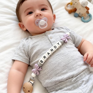 Op de afbeelding: Een baby in een grijze outfit met een fopspeen. Een fopspeenclip met de naam "BRANDY" is bevestigd aan de kleding van de baby. Een houten en siliconen bijtspeeltje staat op de achtergrond.