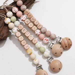 May include: Three baby pacifier clips with wooden beads spelling out names, floral and patterned beads, and wooden clips. The clips are in shades of pink, green, and beige, with a natural wood accent.
